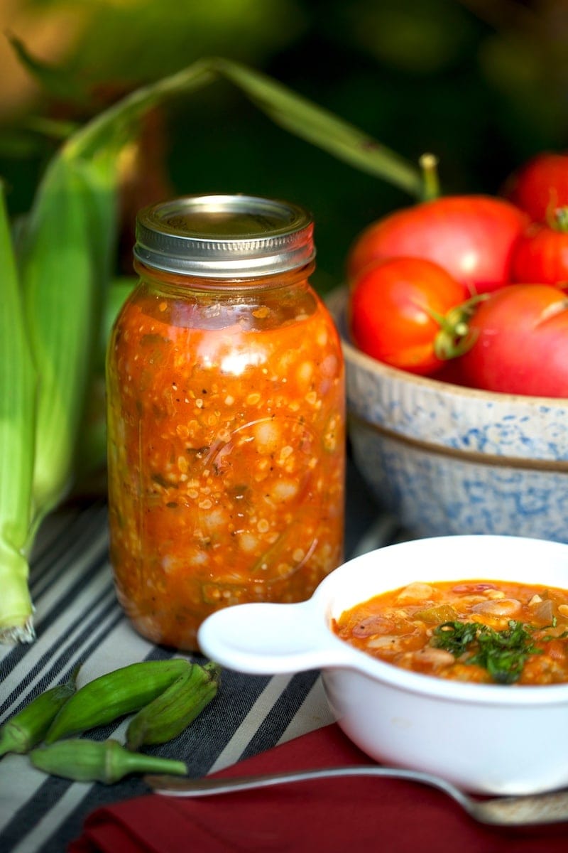 Canned jar of homemade vegetable soup, bowl of tomatoes, and serving of soup in white cup
