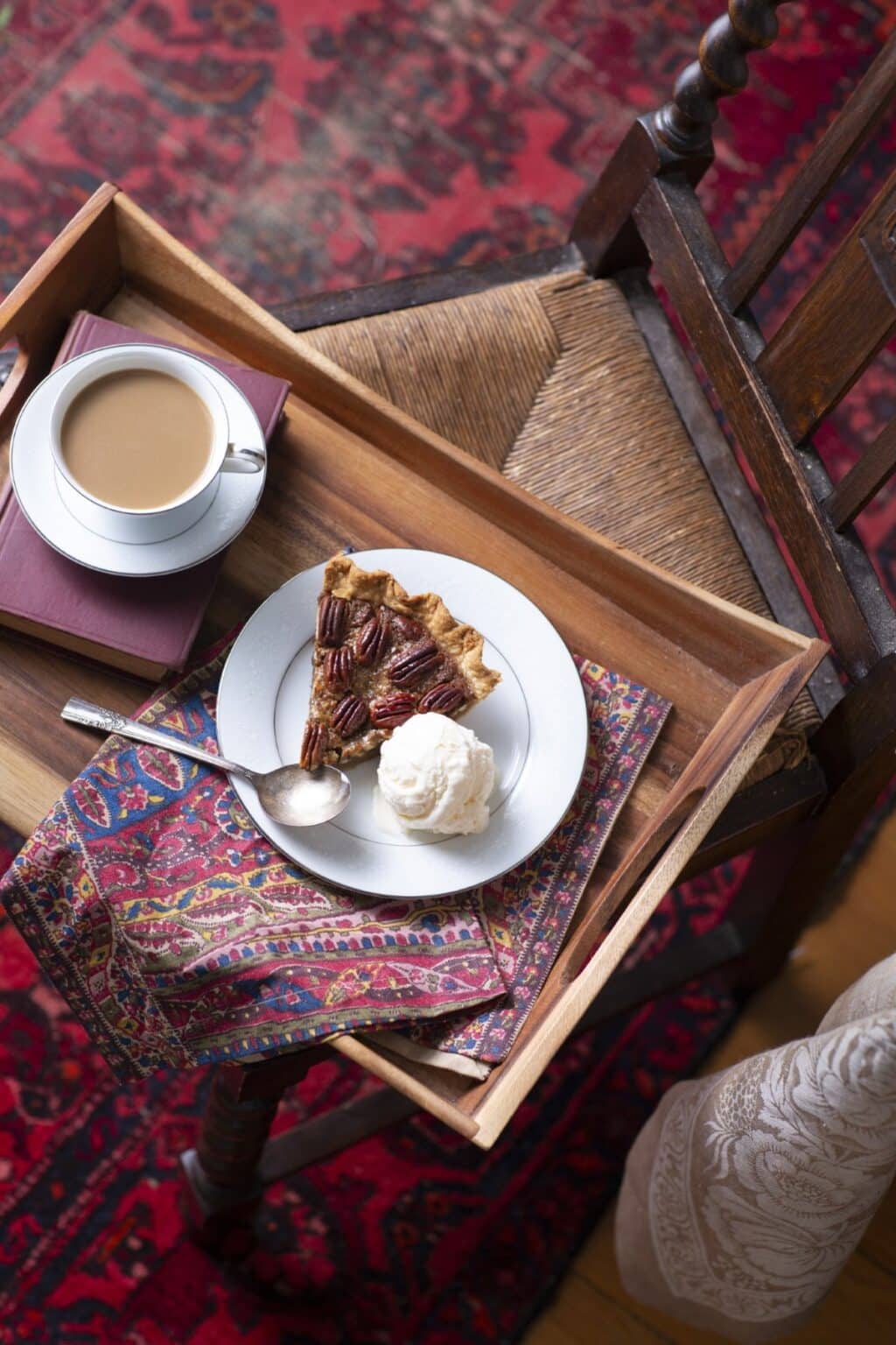 slice of pecan pie with vanilla ice cream on a wooden tray with a cup of coffee and a red-and-blue paisley floral napkin