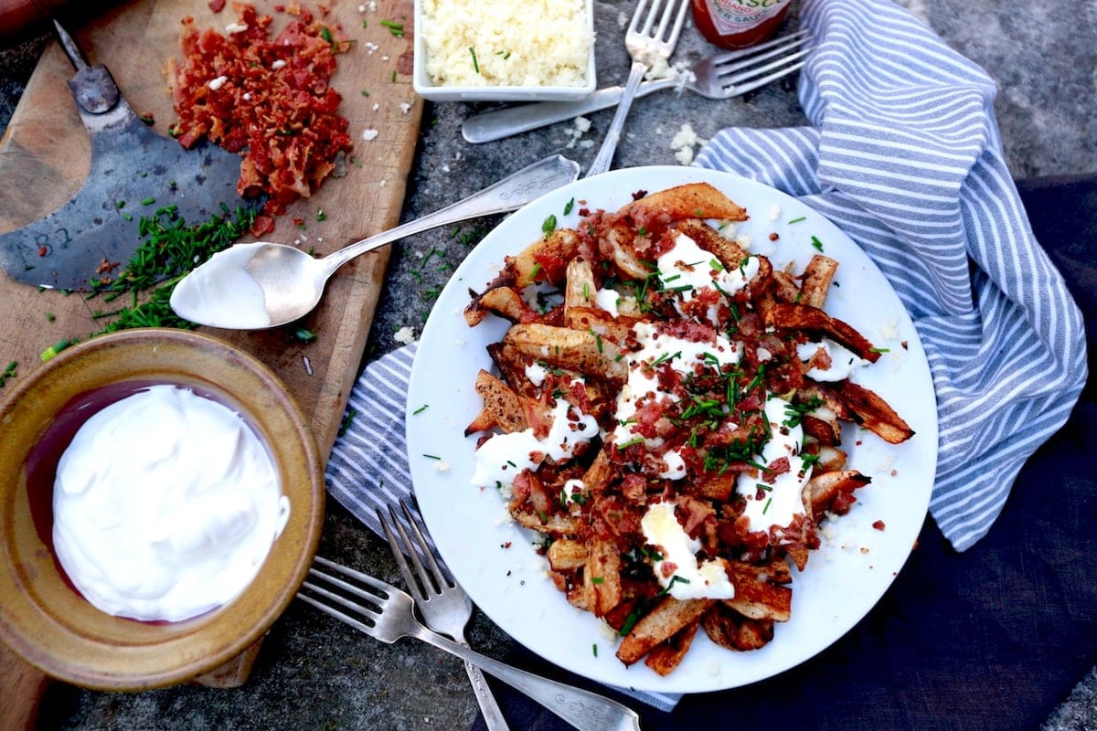 Turnip Root - Loaded Baked Turnip Fries on cement table with bacon, chives, sour cream on a white plate with a striped napkin under it