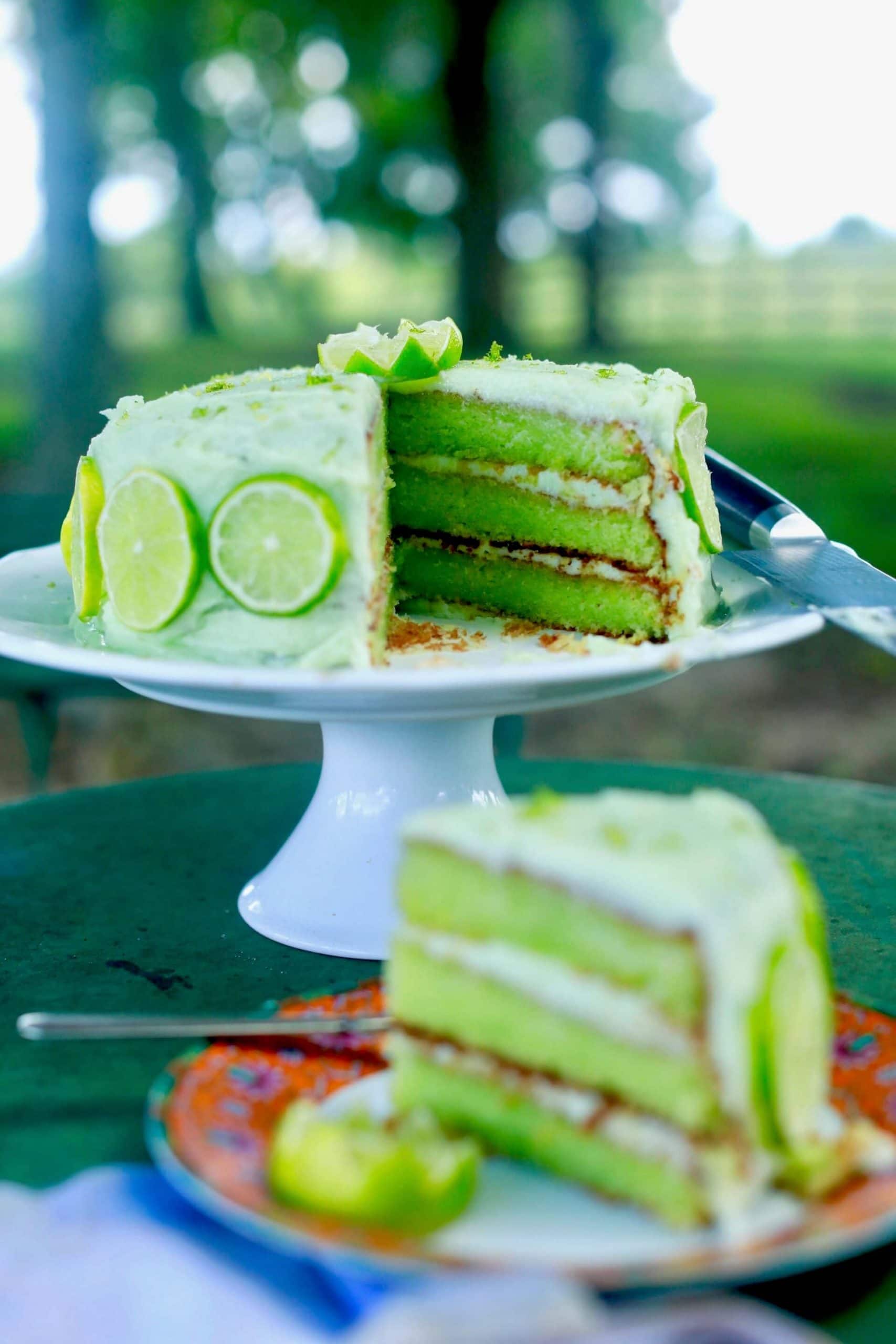 3 layers of green key lime cake with key lime icing with limes around the sides on a white cake stand outside on a table