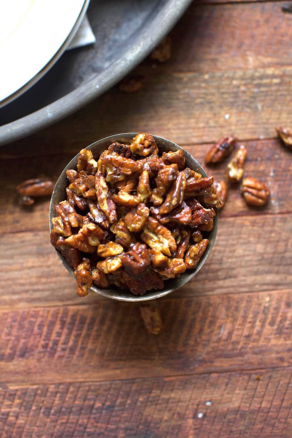 candied pecans served in a small cup resting on rustic wooden table with a few pecans spilled over onto table