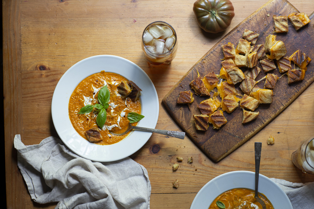 horizontal photo of bowl of roasted tomato soup and glass of tea next to wooden cutting board with cubes of grilled cheese sandwich croutons
