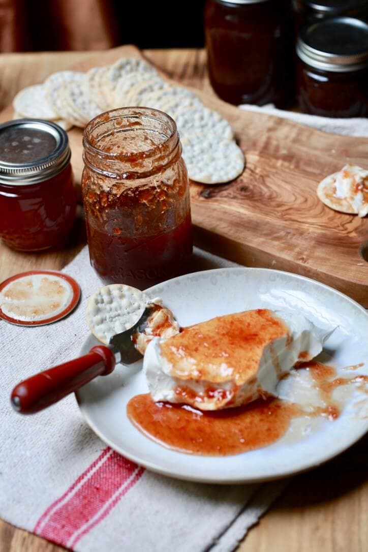 Photo of a table spread including homemade tomato jelly in jars - a plate with tomato jelly spread over a block of cream cheese and a cracker - in background, a cutting board with crackers. Recipe by Stacy Lyn Harris