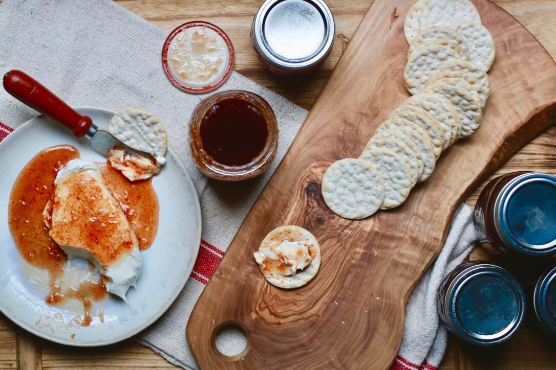 snack spread with jar of tomato jelly, cream cheese and crackers with topping of jelly on white plate, and more crackers on natural wood cutting board