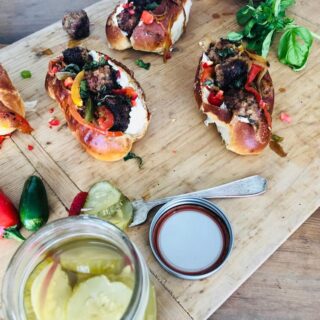 A picture of Venison Meatball Rolls with Horseradish mayonnaise being prepared on a wooden slab, with pickles. Recipe by Stacy Lyn Harris