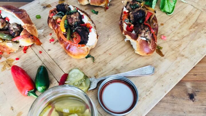 A picture of Venison Meatball Rolls with Horseradish mayonnaise being prepared on a wooden slab, with pickles. Recipe by Stacy Lyn Harris