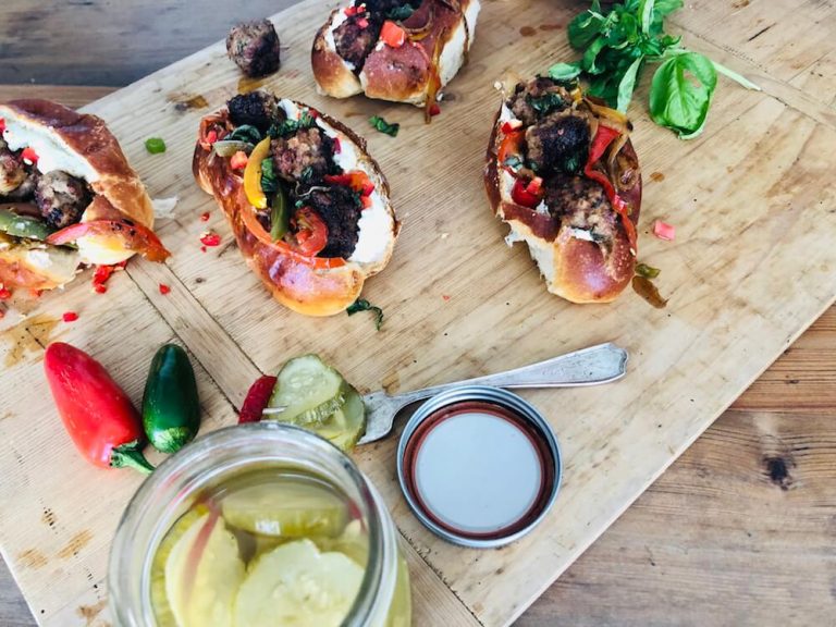 A picture of Venison Meatball Rolls with Horseradish mayonnaise being prepared on a wooden slab, with pickles. Recipe by Stacy Lyn Harris