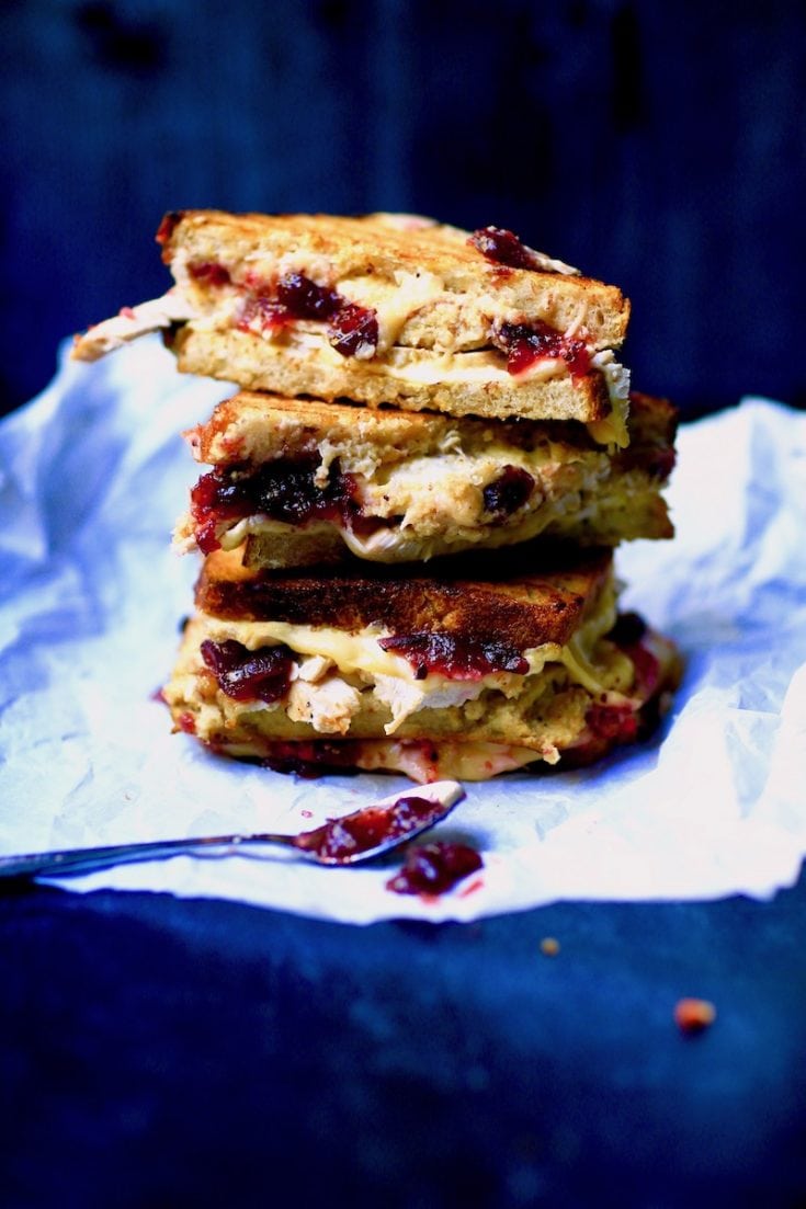 3 turkey and dressing sandwiches halved showing the cranberry sauce, melted cheeses on parchment paper with spoon having been in cranberry sauce