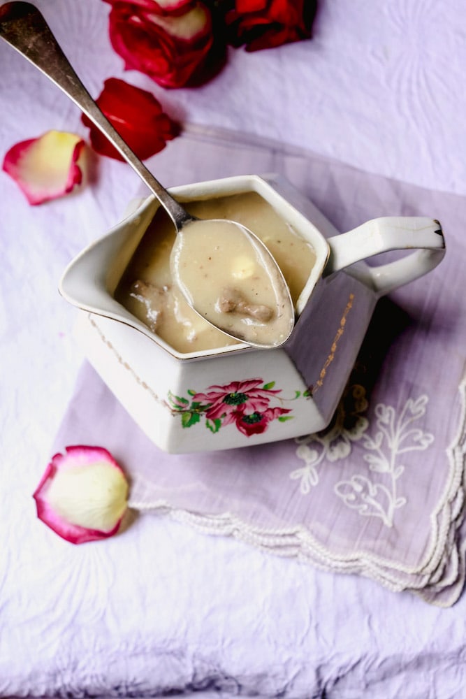 Giblet Gravy in a rose stenciled serving dish with spoon of gravy laying on top and rose petals surrounding it on lavender tablecloth