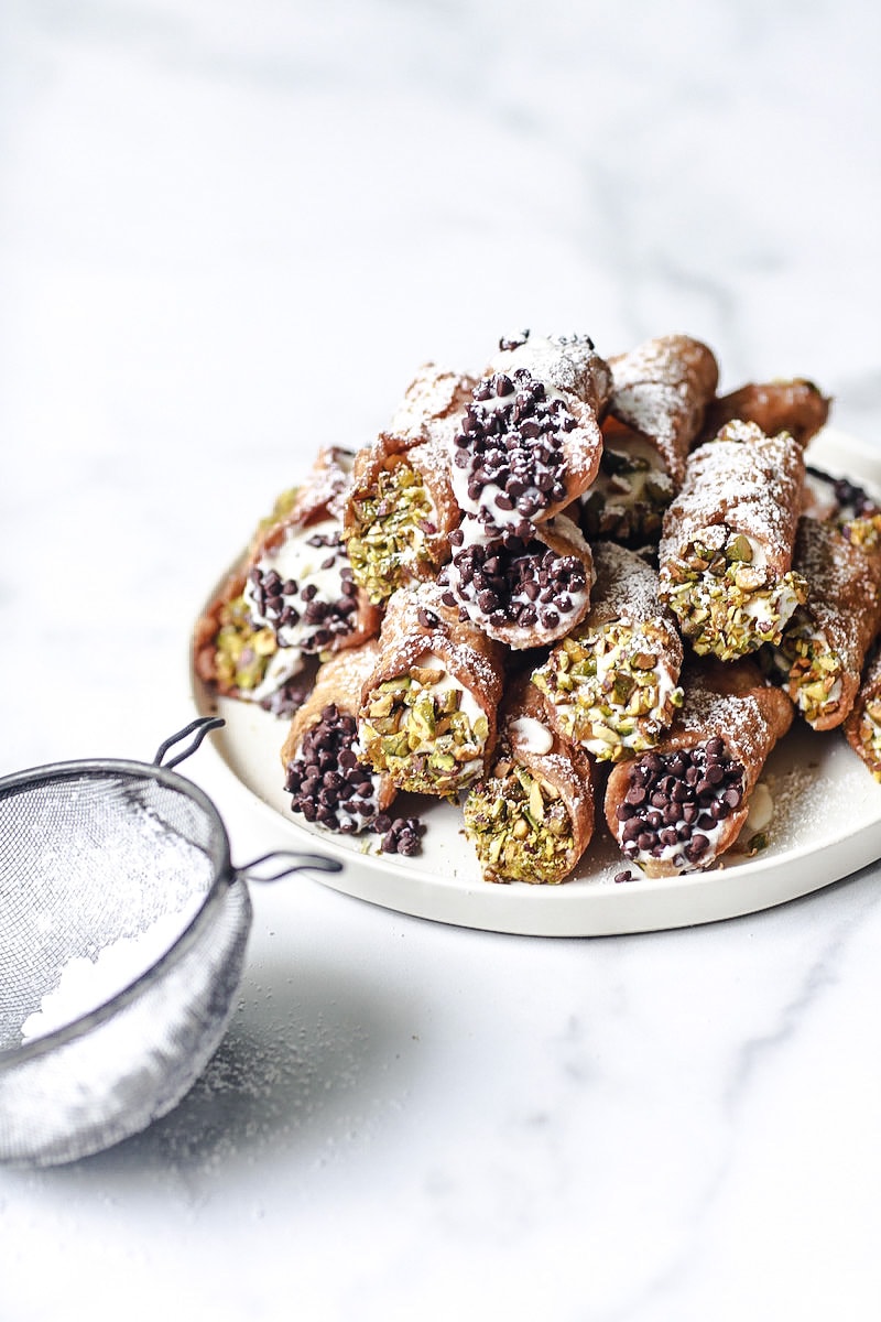 Canolli served on a white plate on white marble with chocolate and pistachio on the sides