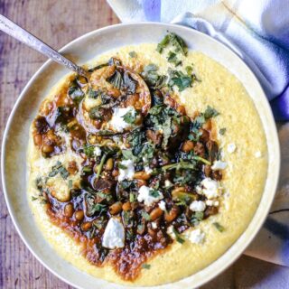 Bowl of Persian Stew with venison, beans, collards, spices and herbs, and topped with goat cheese in a bowl of cheesy grits with folded napkin on wooden table