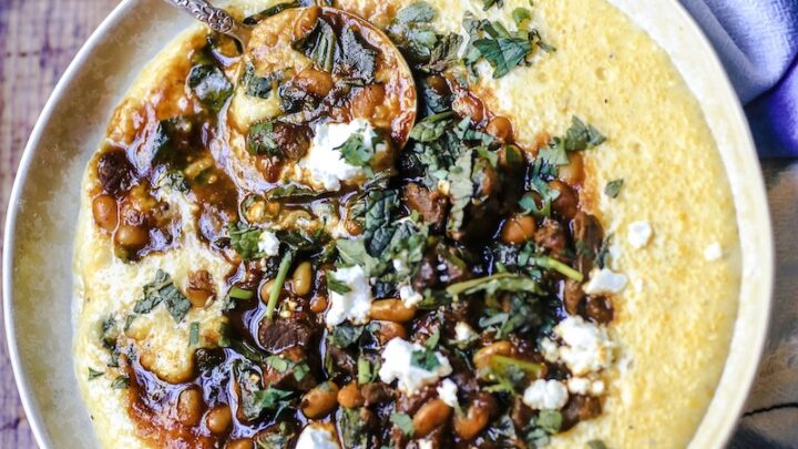 Bowl of Persian Stew with venison, beans, collards, spices and herbs, and topped with goat cheese in a bowl of cheesy grits with folded napkin on wooden table