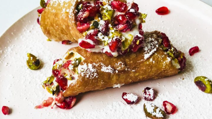 Cannoli on a pink plate with a pomegranate in the background and pomegranate and pistachios