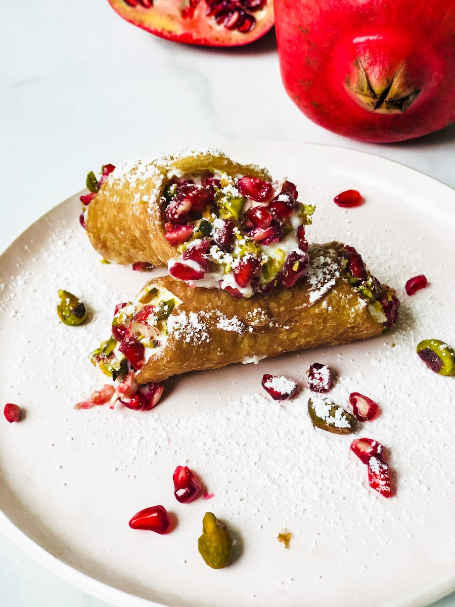 Cannoli on a pink plate with a pomegranate in the background and pomegranate and pistachios