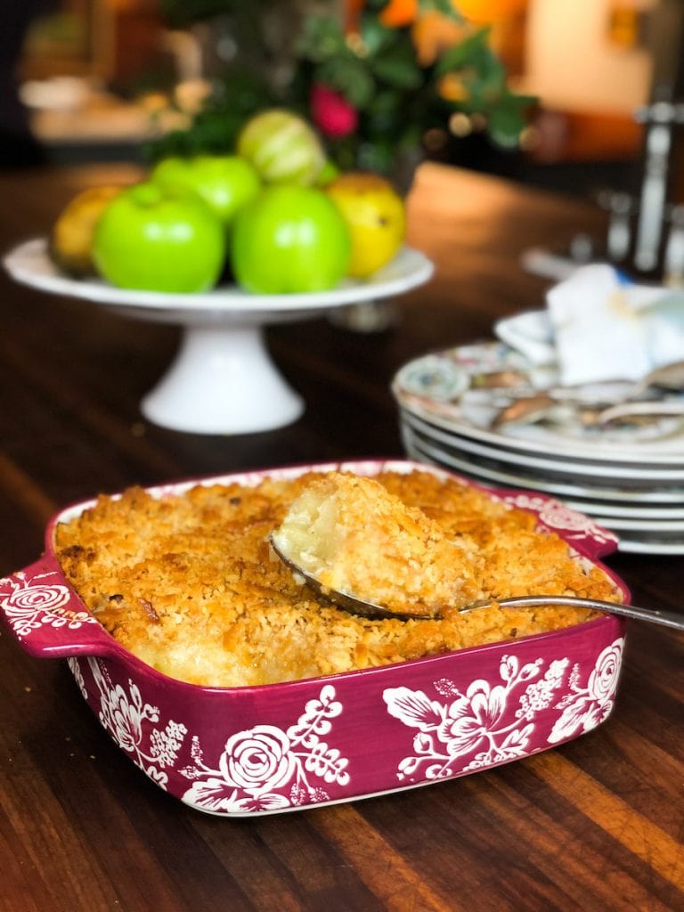 Pineapple Casserole with Buttery Tollhouse Crackers in Red Casserole Dish with White Flowers and cake stand with apples in background
