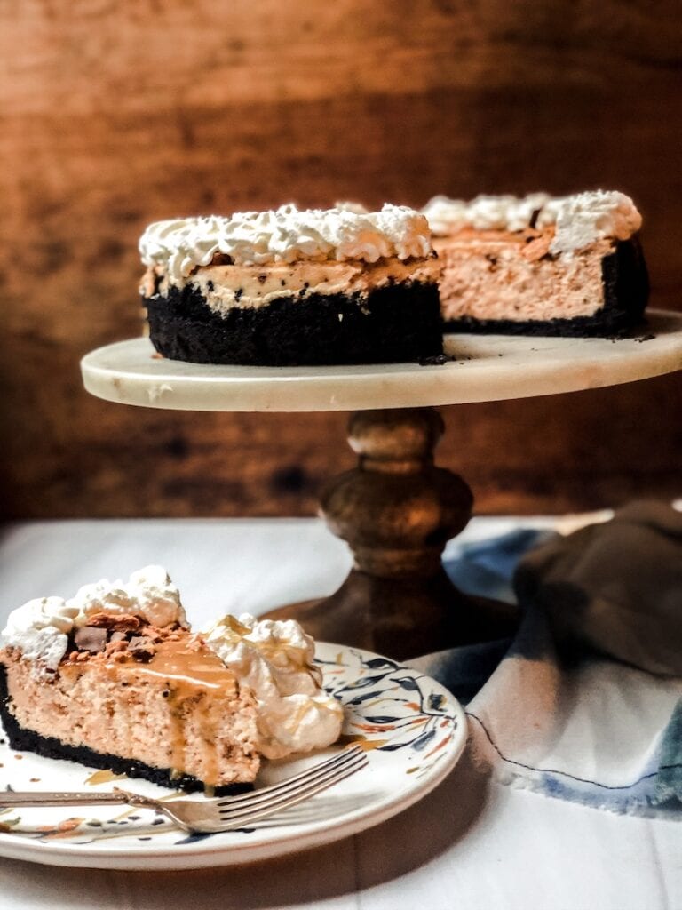 Best Butterfinger Cheesecake on cake stand with plate under it on white surface with a silver fork