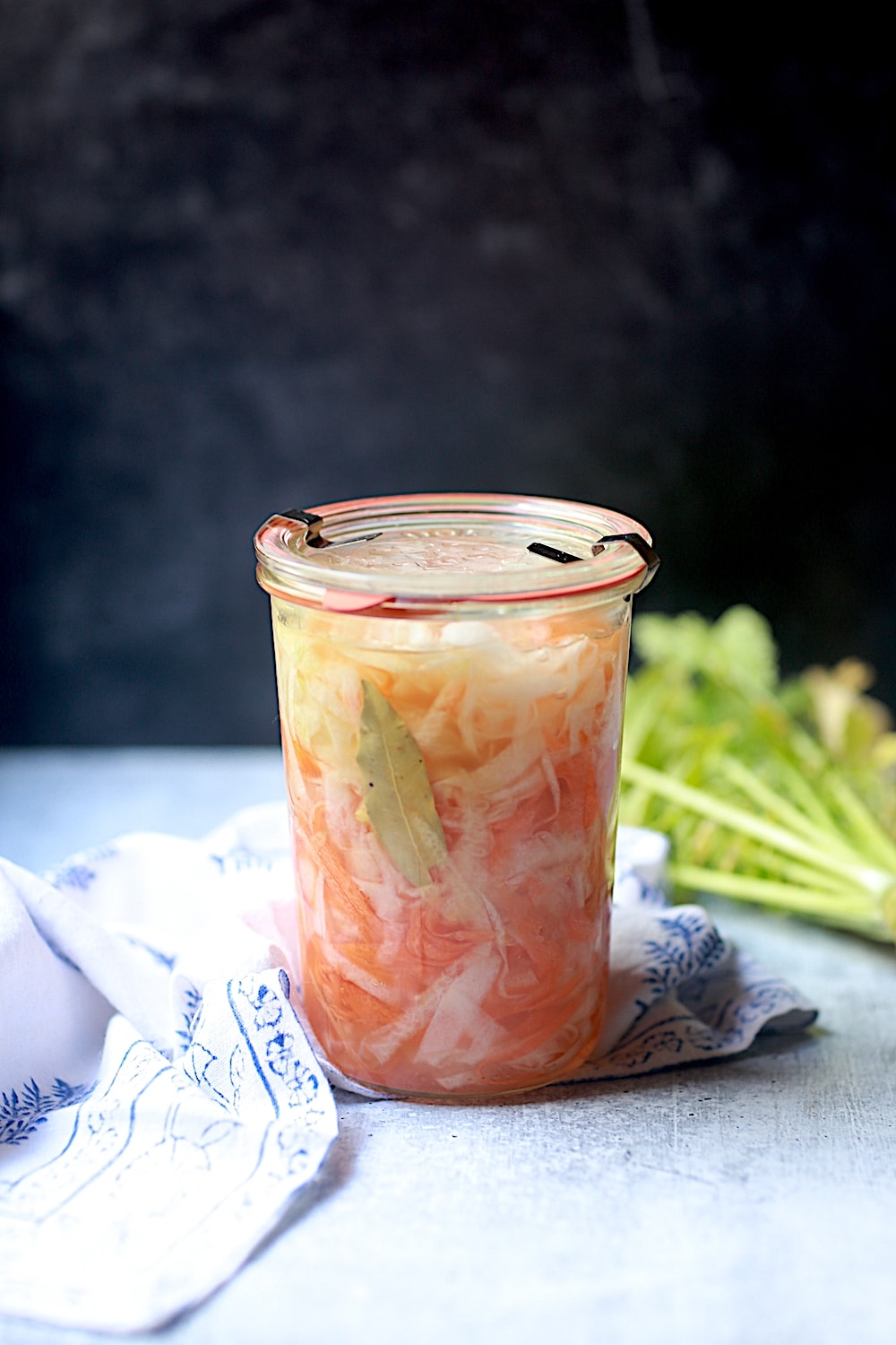 Jar of Pickled Daikon and Carrots with a blue and white napkin and radish leaves behind the jar