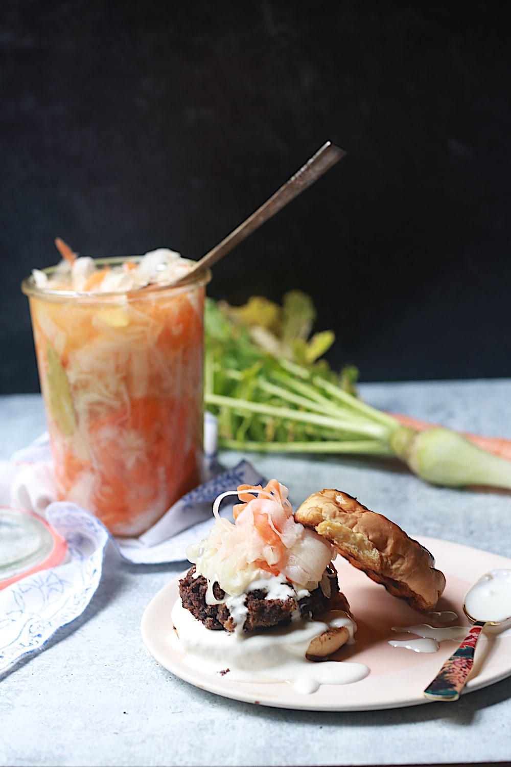 Burger topped with Pickled daikon and carrots radishes on pink plate with the pickled vegetables and radishes in background