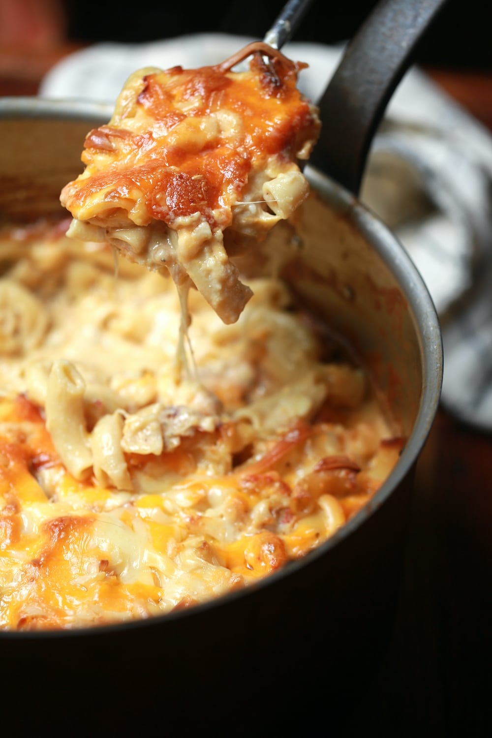 Crawfish mac and cheese with spoon showing the gooey cheese in copper pot with steel handle and napkin behind it.
