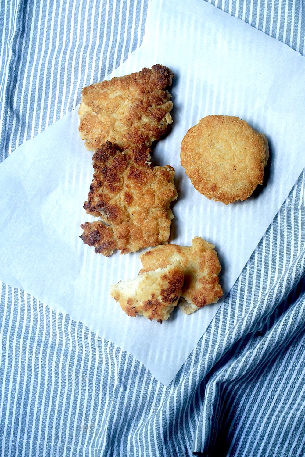 Fried Cornbread, some with more rustic and rough edges and some perfectly round, on a piece of parchment paper on top of a blue and white striped linen.