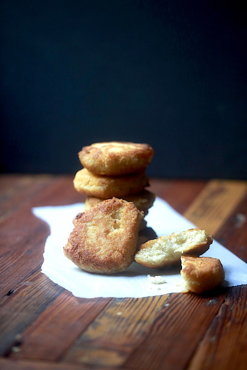 Stacked Fried Cornbread with a piece of hot water cornbread propped up showing the crispy outside and one cut open showing the creamy inside on wood with a black background.