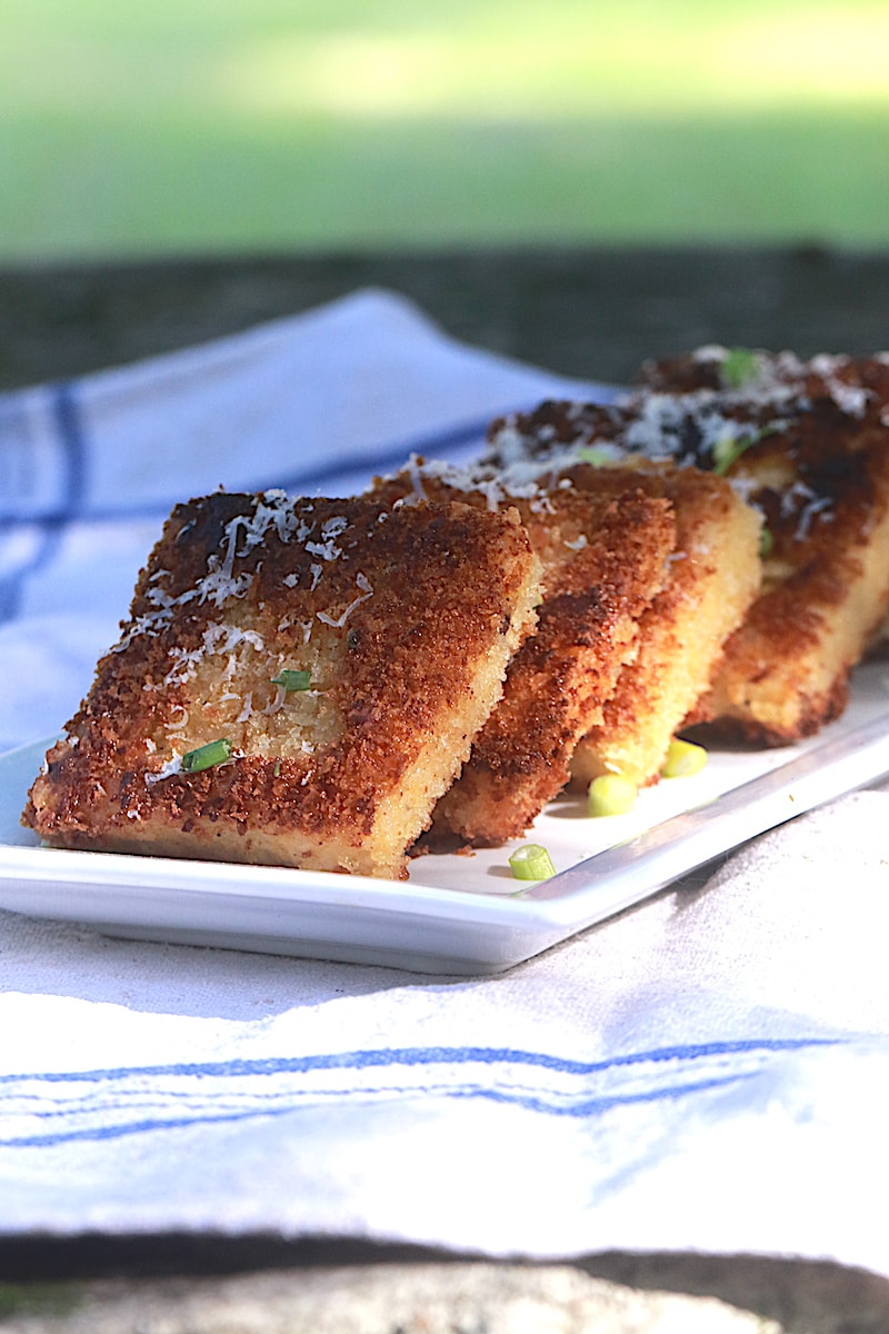 Fried Grits Cakes with Parmesan and scallions stacked on top of a rectangular white plate on top of a blue and white napkin