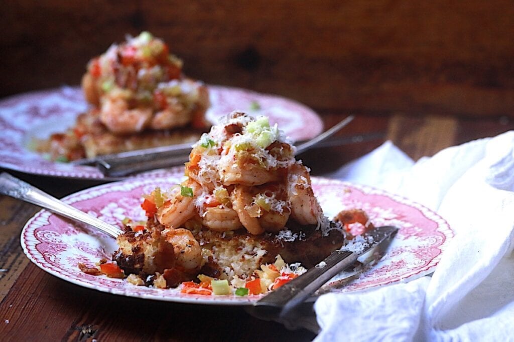 Shrimp piled on top of a fried cheesy grits cake with chopped bell pepper and onions on pink and white plates with a spoon and knife.