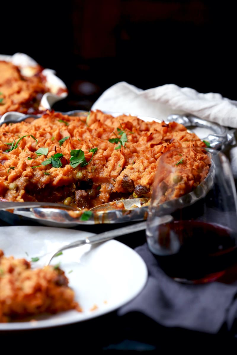 Deer meat shepherd's pie with sweet potatoes over the top in a silver serving dish. A piece of venison shepherd's pie Is in the foreground on a white dish.