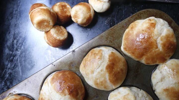 Overhead shot of dinner rolls in pan and some on a black surface with a knife spooning butter on top.