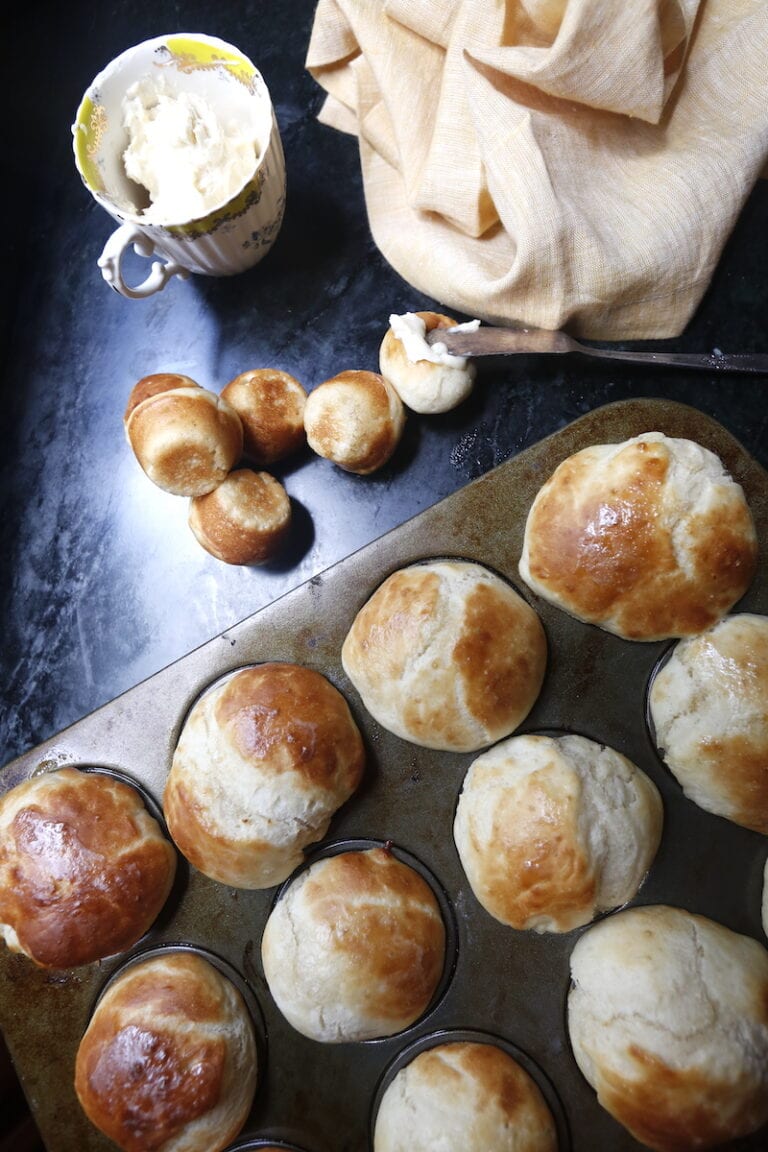 Overhead shot of dinner rolls in pan and some on a black surface with a knife spooning butter on top.