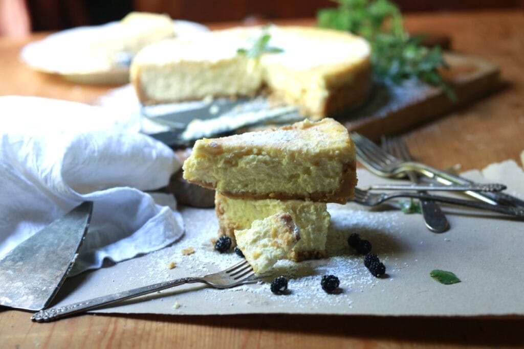 Stacked slices of Italian cheesecake on paper with forks and knife in front of freshly baked cake