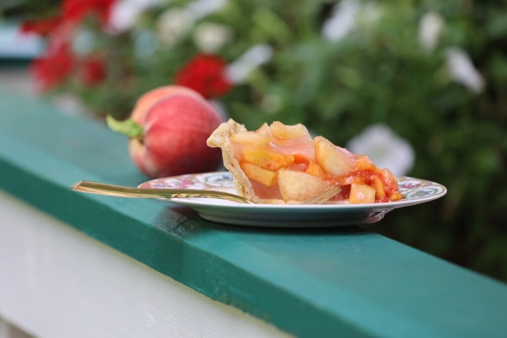 slice of peach pie on china plate with fresh peach balanced on green porch railing