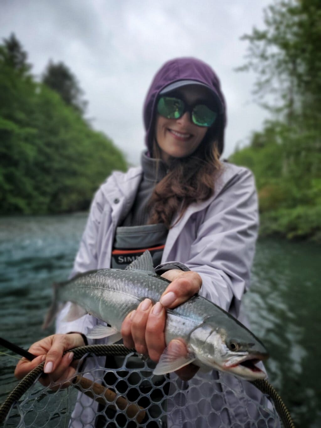 Stacy Lyn smiling in fly fishing gear in Alaska creek, holding fishing net and large fish