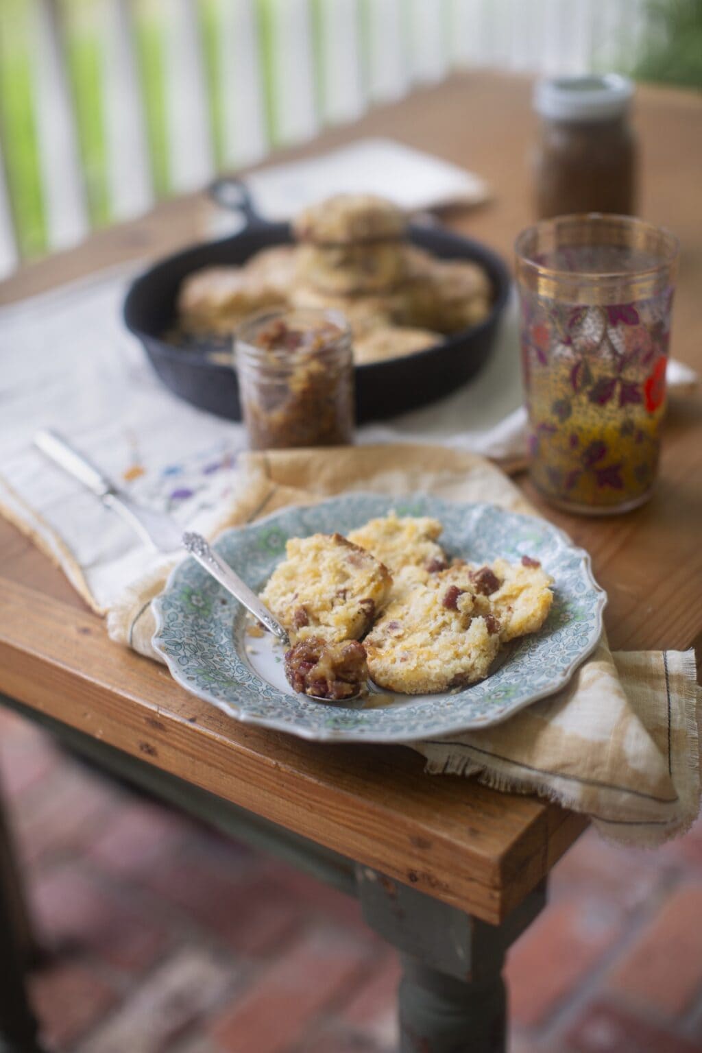 On wooden table, a blue china plate in foreground with sliced cheesy biscuits and a spoon with dollop of bacon jam; glass with juice, glass jar of jam, and biscuit pan in background.