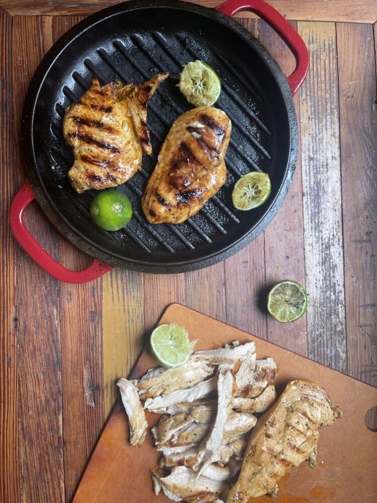 foreground is cutting board with sliced skinless chicken breast, background is grilled chicken breasts in black pan