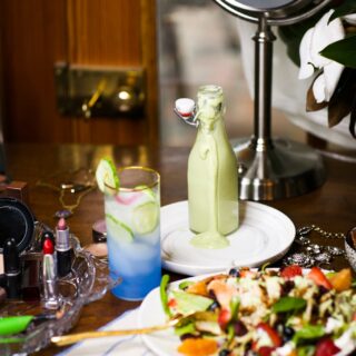 avocado dressing, green goddess style, in glass bottle behind white plate full of power salad with chicken and a blue beverage with ice. Salad is next to a tray with lipsticks.