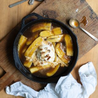 bananas foster with vanilla ice cream in cast iron skillet on wooden cutting board, next to metal spoons and crumpled white napkin