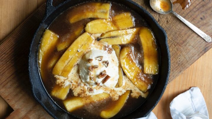 bananas foster with vanilla ice cream in cast iron skillet on wooden cutting board, next to metal spoons and crumpled white napkin