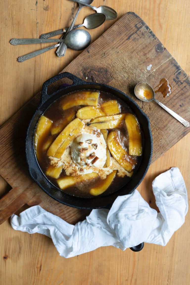 bananas foster with vanilla ice cream in cast iron skillet on wooden cutting board, next to metal spoons and crumpled white napkin