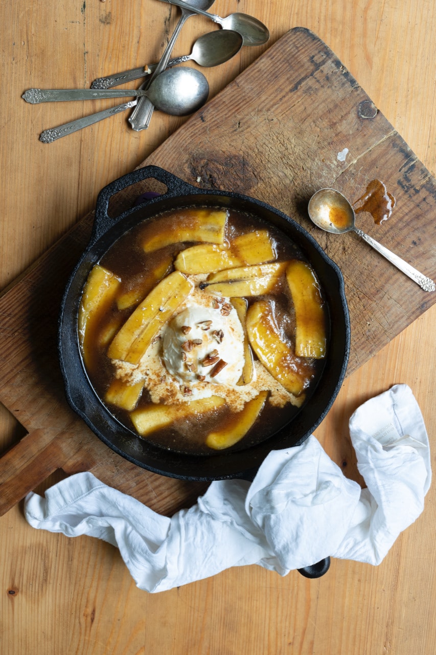 bananas foster with vanilla ice cream in cast iron skillet on wooden cutting board, next to metal spoons and crumpled white napkin