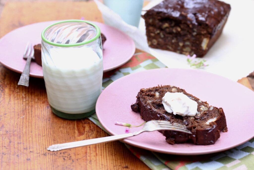 slice of chocolate chocolate-chip banana bread with butter on pink plate next to glass of milk, loaf in background