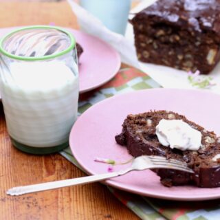slice of chocolate chocolate-chip banana bread with butter on pink plate next to glass of milk, loaf in background