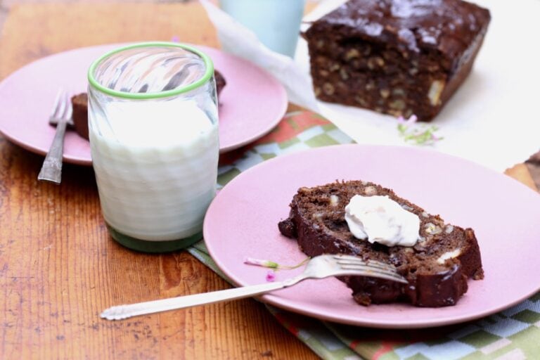 slice of chocolate chocolate-chip banana bread with butter on pink plate next to glass of milk, loaf in background