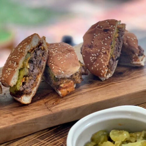 Smash burgers on bun, sliced in half and resting on wooden cutting board with side of pickles, from Stacy Lyn's recipes for Father's Day