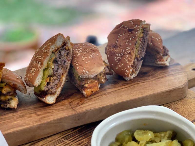 Smash burgers on bun, sliced in half and resting on wooden cutting board with side of pickles, from Stacy Lyn's recipes for Father's Day