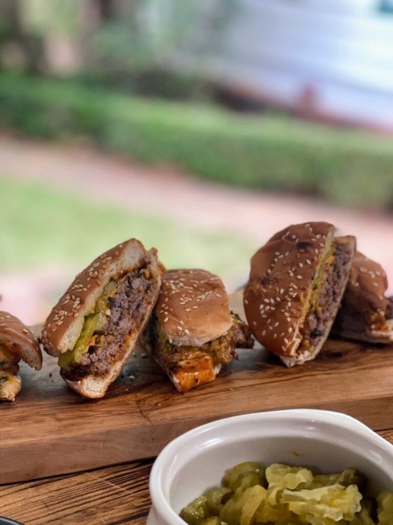 Smash burgers on bun, cut in half and arranged on cutting board behind a bowl of pickle slices