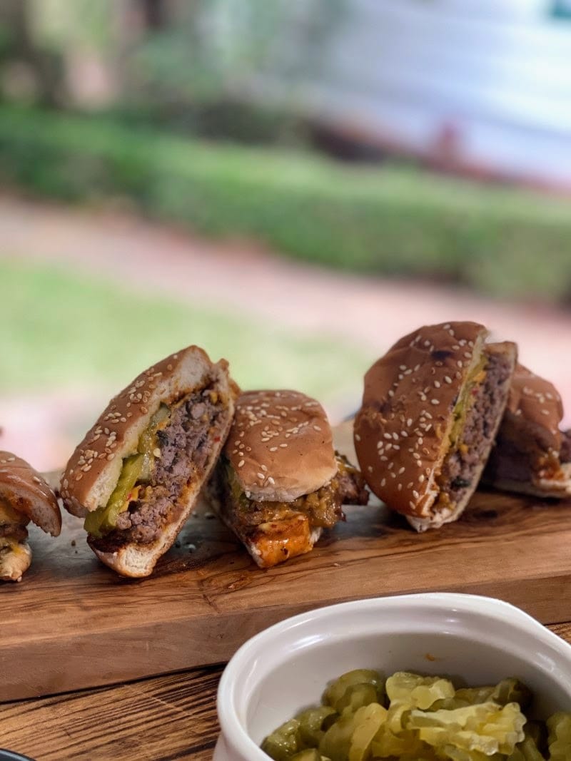 Smash burgers on bun, cut in half and arranged on cutting board behind a bowl of pickle slices