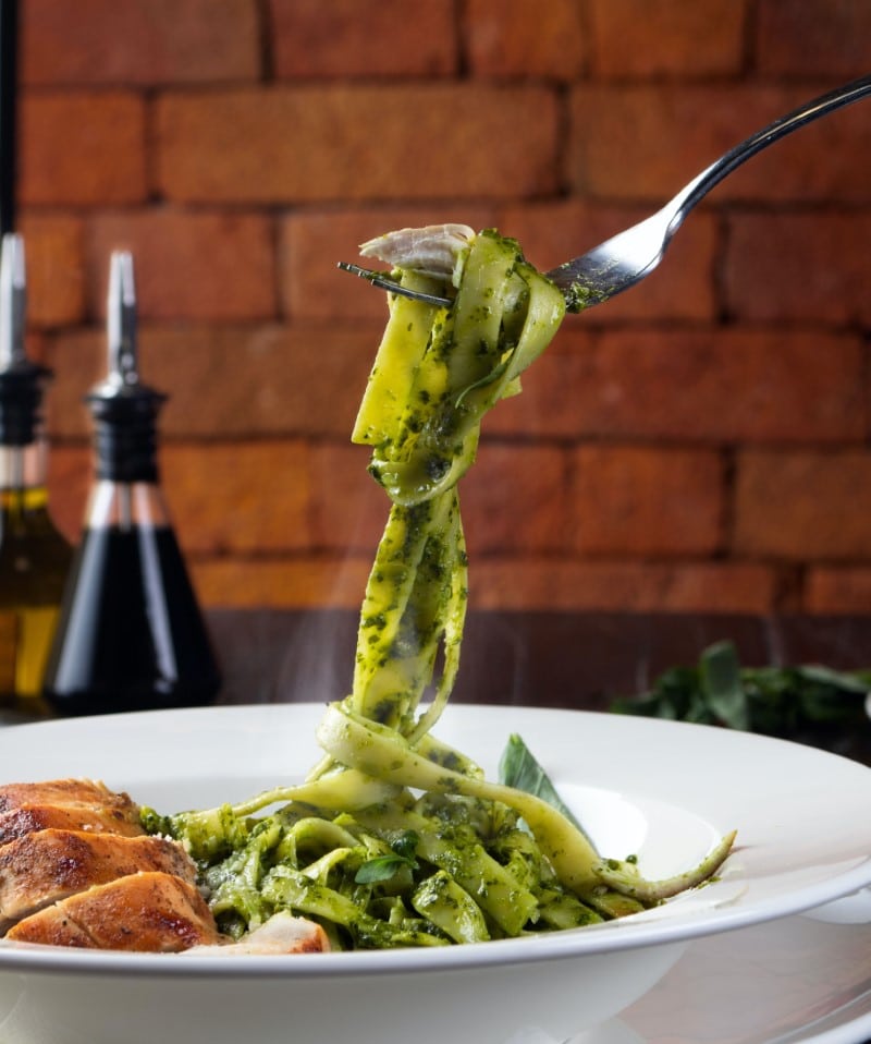 basil pesto on pasta in white bowl with a fork twirling the pasta, chicken breast on the side, brick wall in background