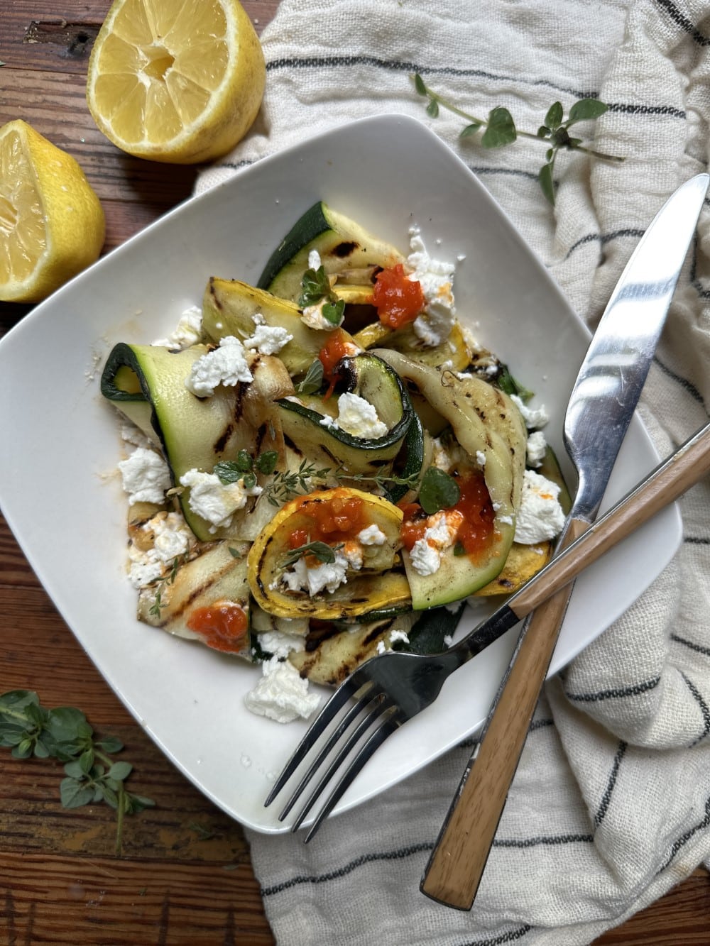 closeup of zucchini salad in white, square shallow bowl with crossed knife and fork, white towel and sliced lemon in background