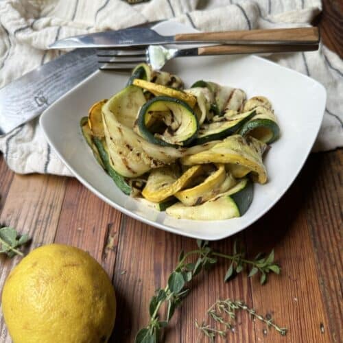 serving of grilled zucchini salad in white, shallow square bowl with fork and knife, lemon and herbs in foreground, and white towl in background on wooden table